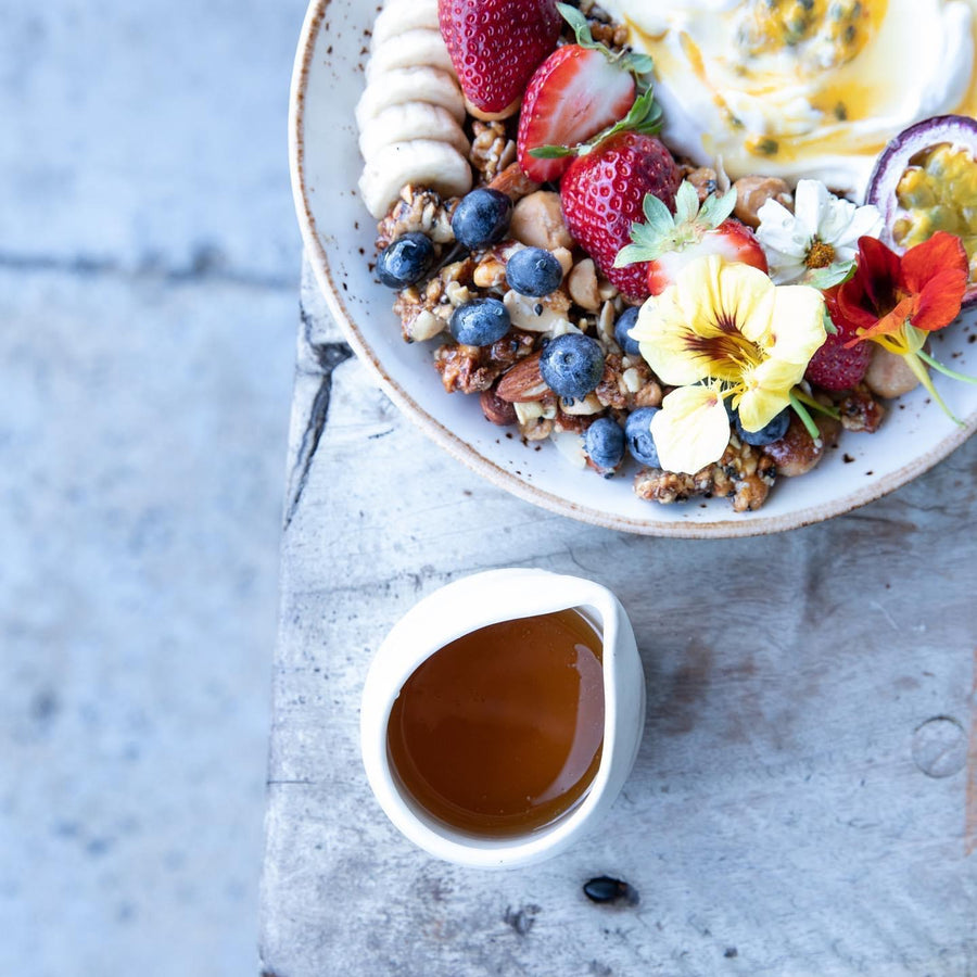 A jug of honey next to bowl of granola with fresh blueberries, strawberries, bananas, yoghurt and passionfruit