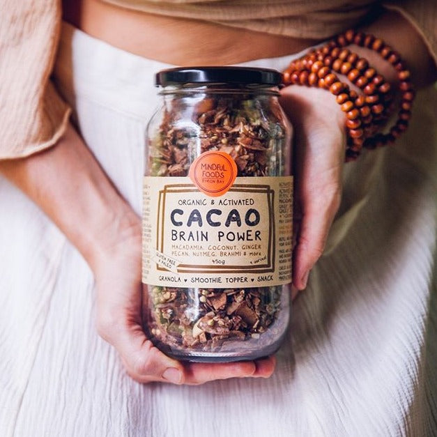 Person's hands holding 450g glass jar of Organic & Activated Cacao Brain Power showing ingredients of macadamia, coconut, ginger, pecan, nutmeg, brahmi & more.  