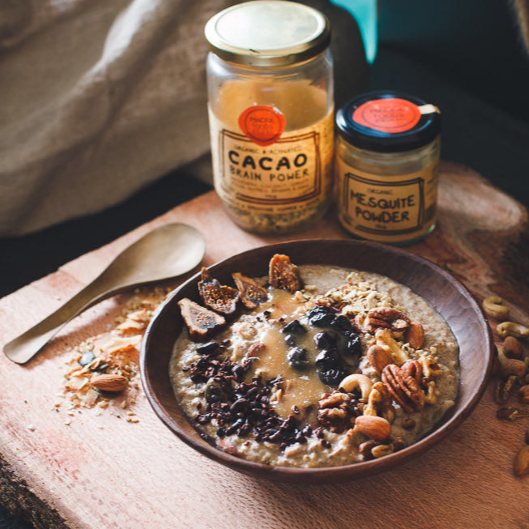 Wooden Bowl of porridge with cacao brain power, fresh figs, pecan nuts and others, fresh berries. Half filled glass jar of cacao brain power next to small jar of mequite powder.  Wooden spoon to left of bowl.  