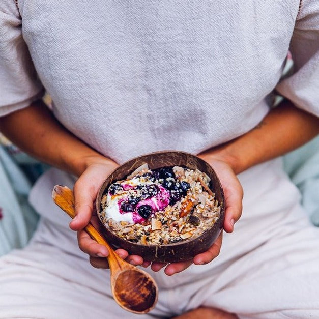 Person holding coconut shell bowl and spoon with golden granola topped with yoghurt and berries