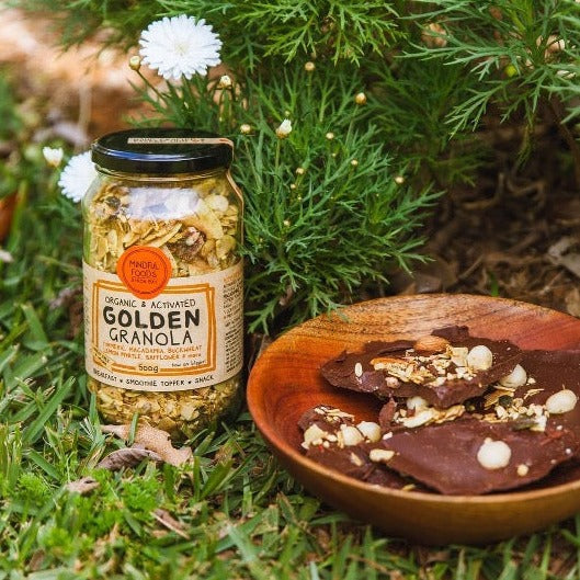 Glass Jar of Organic & Activated Golden Granola in the garden next to wooden bowl with chocolate clusters made with golden granola