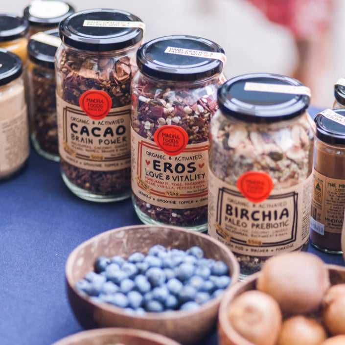 3 glass jars of Mindful Foods granolas displayed on table - organic & activated cacao brain power, Eros Love & Vitality and the Birchia paleo prebiotic granola.  Blueberries and kiwi fruit in bowls in the foreground