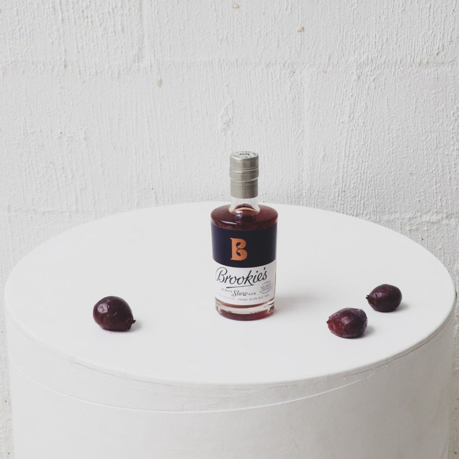 200ml bottle of Brookie's Slow Gin with some Davidson Plums around it on a white round  table.